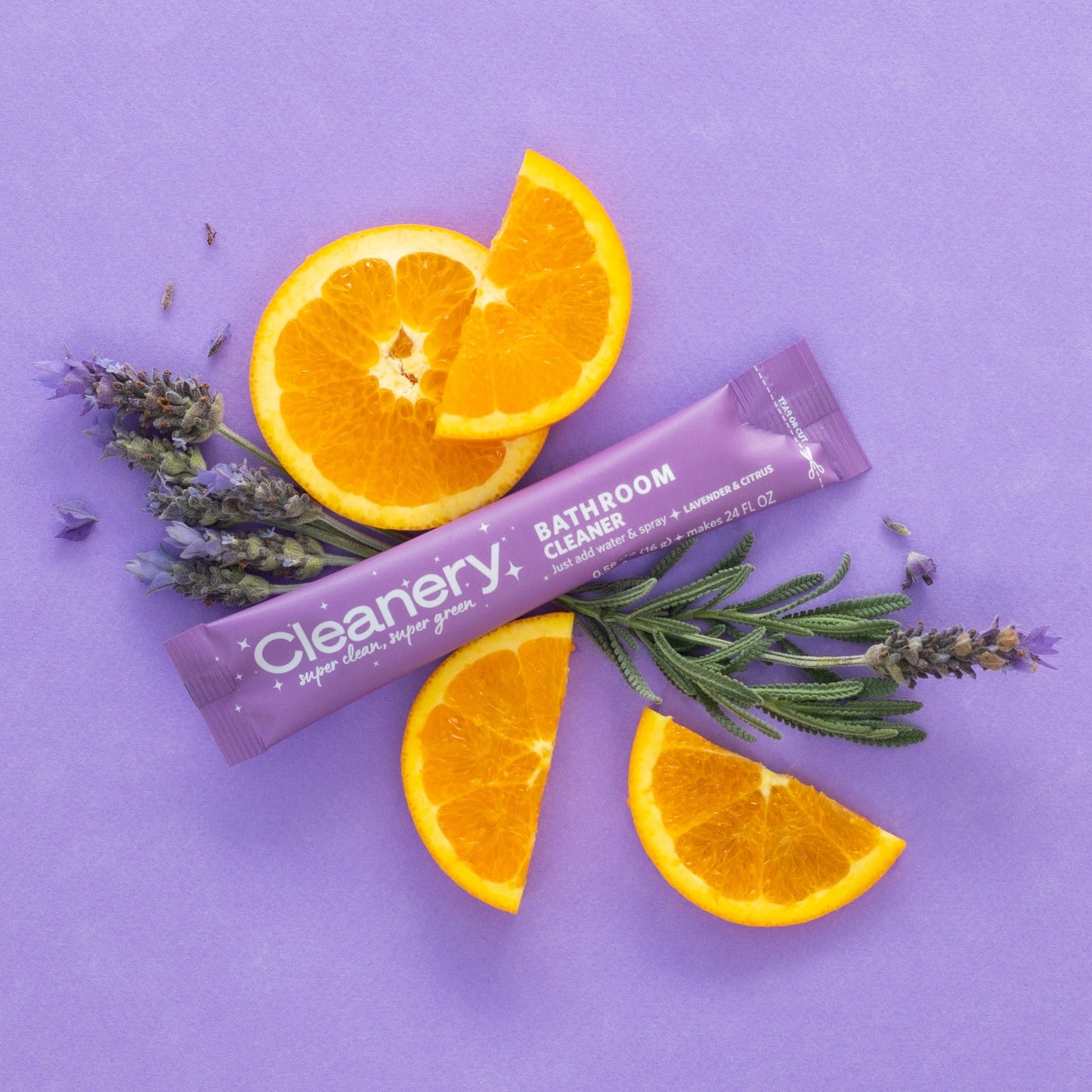 A sachet of Cleanery bathroom cleaning spray with lavender and citrus elements, displayed alongside orange slices and lavender sprigs on a purple background.