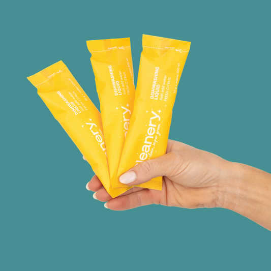 Hand holding three yellow packets of Cleanery on a teal background