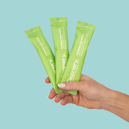 Hand holding three green packets of Cleanery on a light blue background