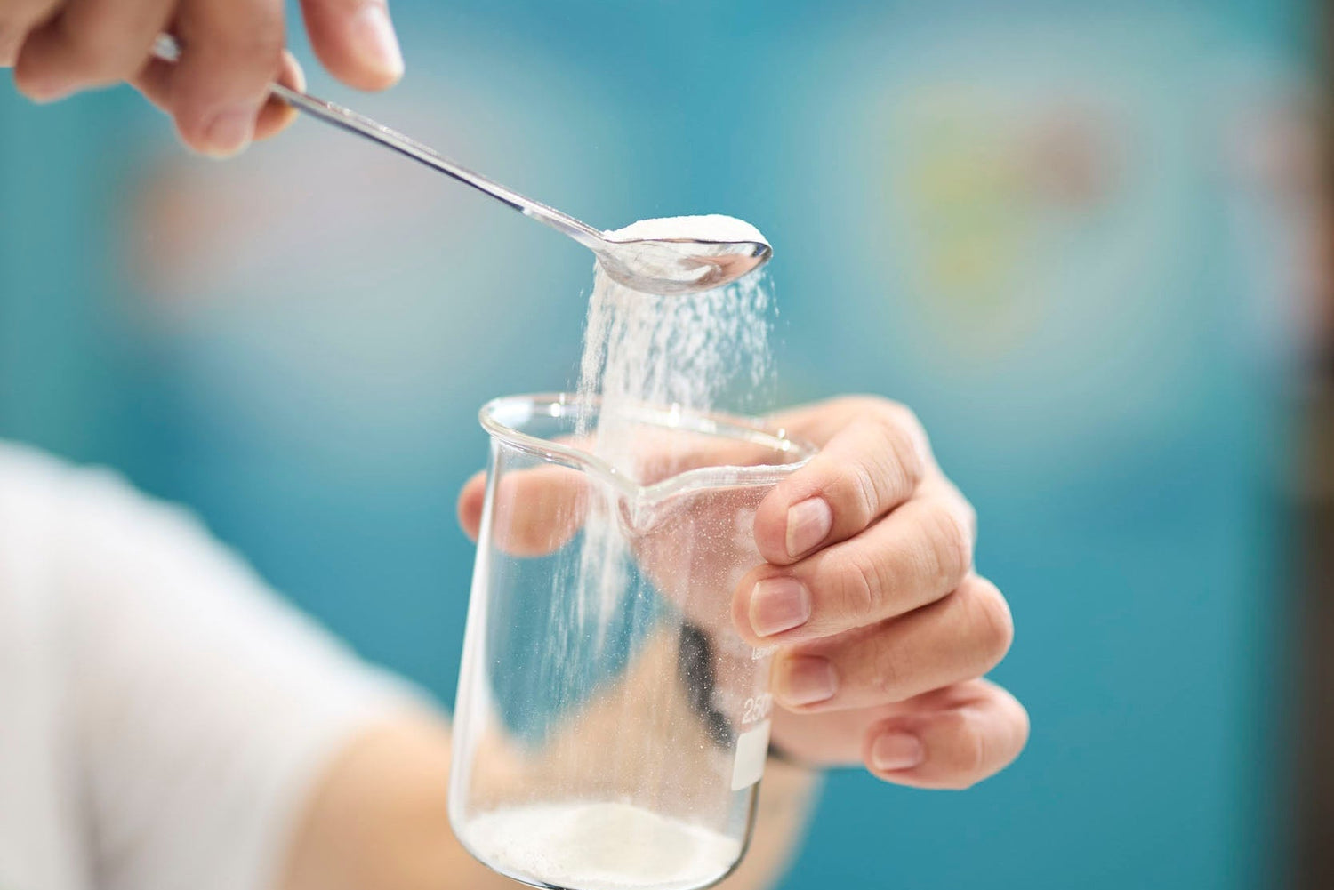 A close-up of a person's hands using a metal sieve to sift white powder into a clear glass beaker, with a blurred blue background.