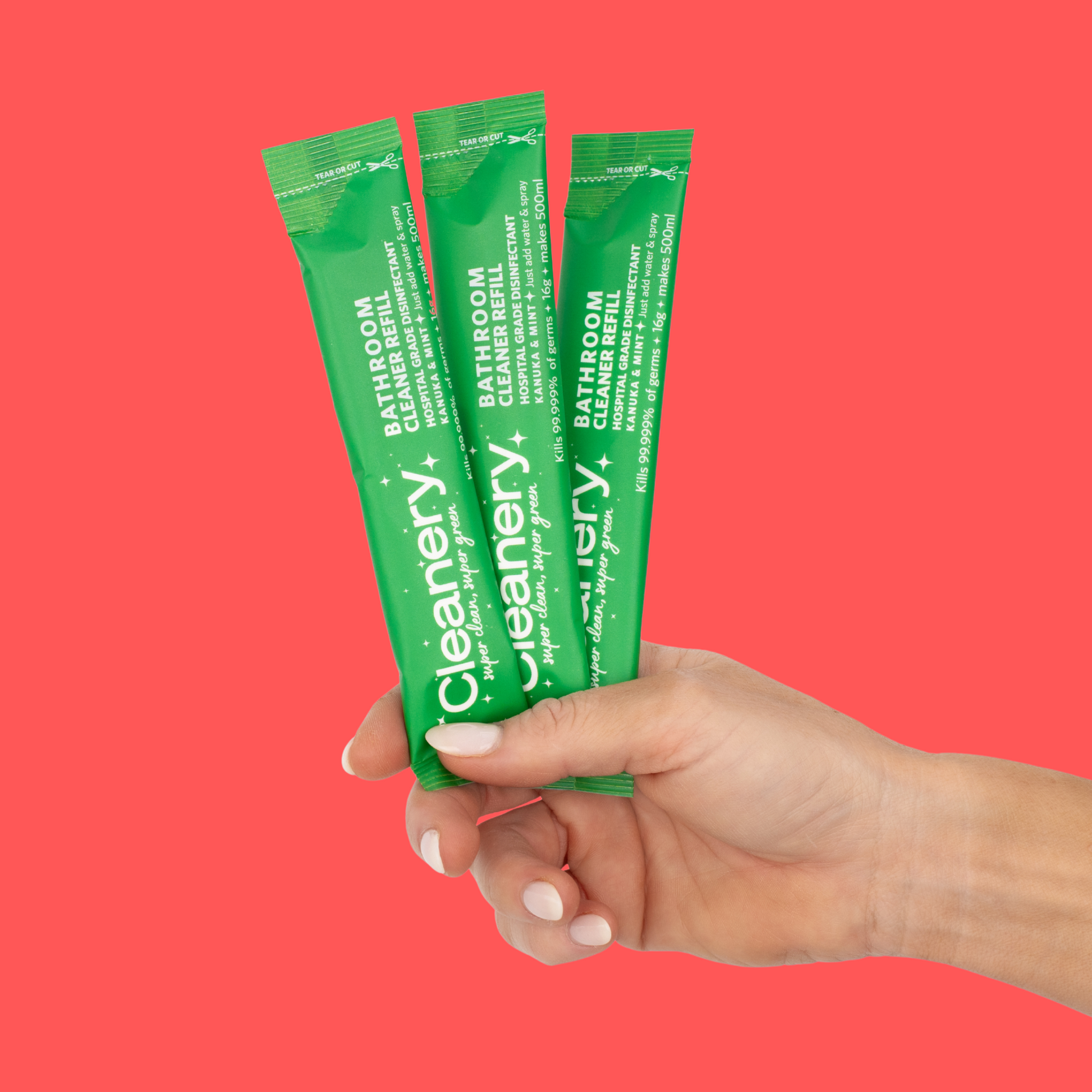 Hand holding three green packets of Cleanery bathroom cleaner against a red background
