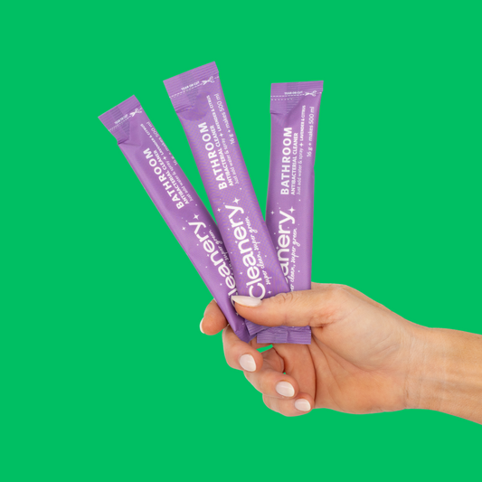 Hand holding three purple 'Cleanery' bathroom cleaning refills against a green background