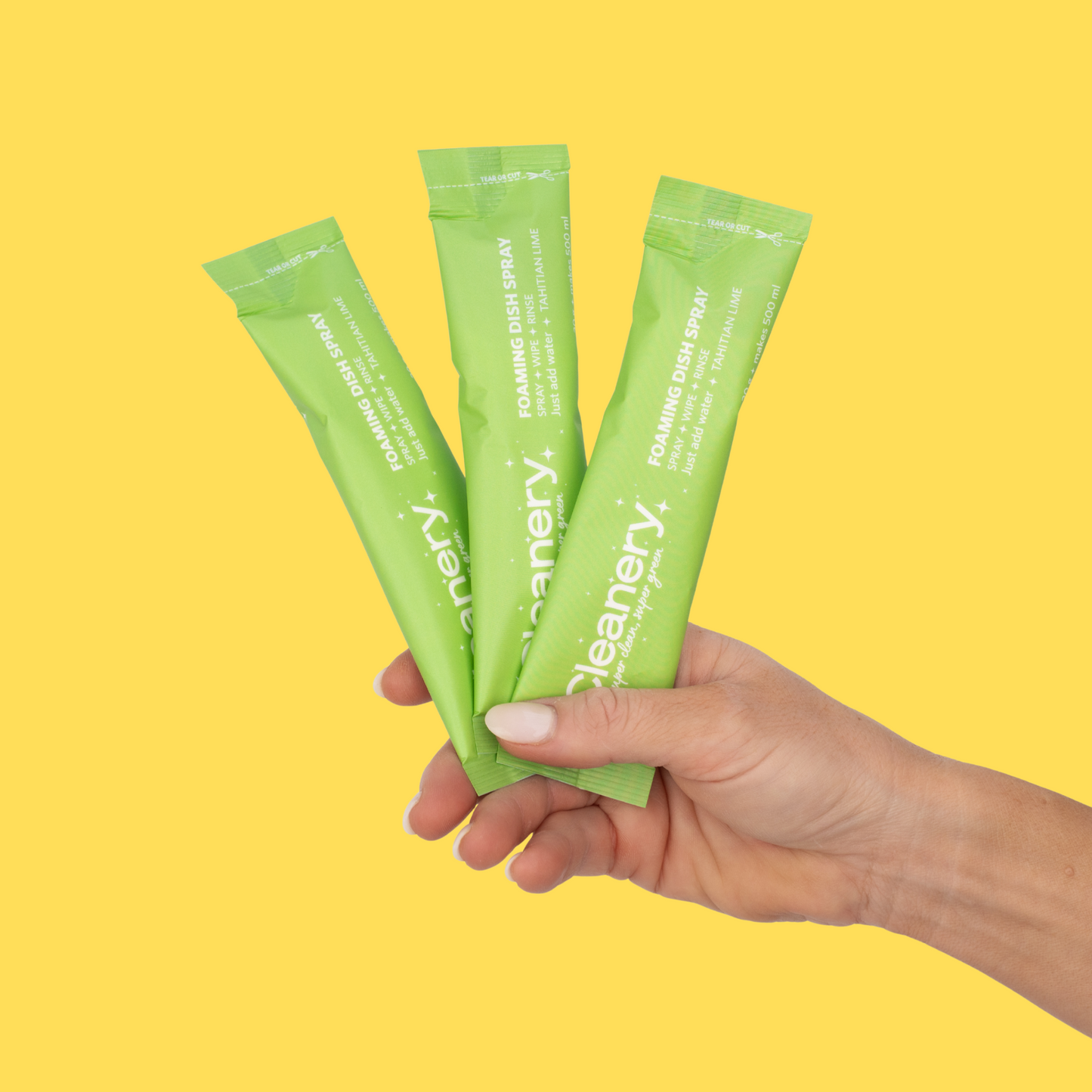 Hand holding three green packets of Cleanery foaming dish spray on a yellow background
