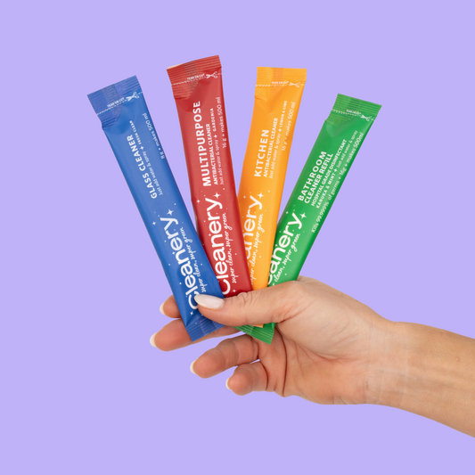 Hand holding four colorful packets of Cleanery cleaning products against a purple background