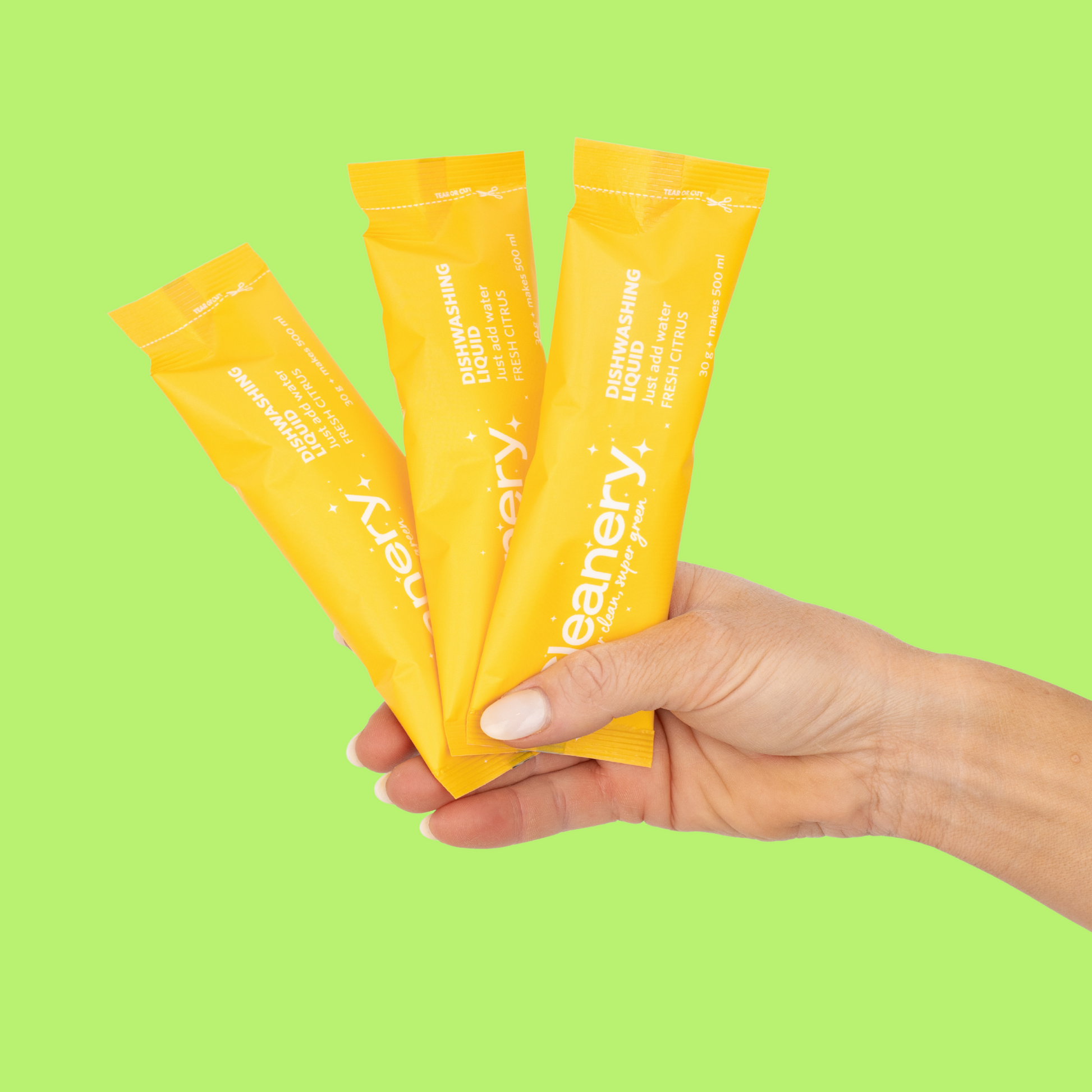 Hand holding three yellow Cleanery dishwashing liquid packets against a green background
