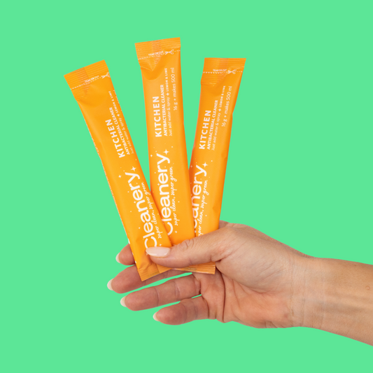 Hand holding three orange packets of Cleanery kitchen cleaner against a green background