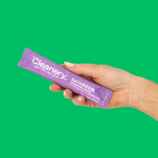 Hand holding a purple 'Cleanery' bathroom cleaning stick against a green background