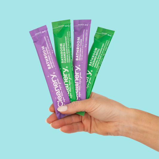A hand holding four Cleanery bathroom refill sachets against a blue background