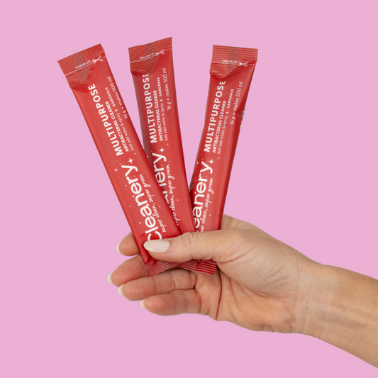 Hand holding three red packets of Cleanery multipurpose cleaning product against a pink background