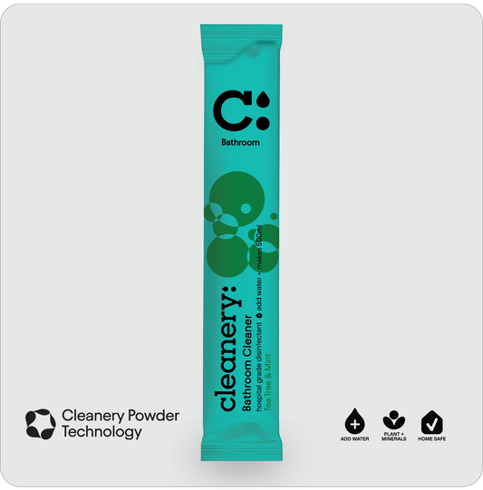 A single teal and green stick pack of Cleanery Bathroom Cleaner centered on a light gray background, with the Cleanery logo and product name clearly visible. Small feature icons appear at the bottom left and bottom right outside the pack.